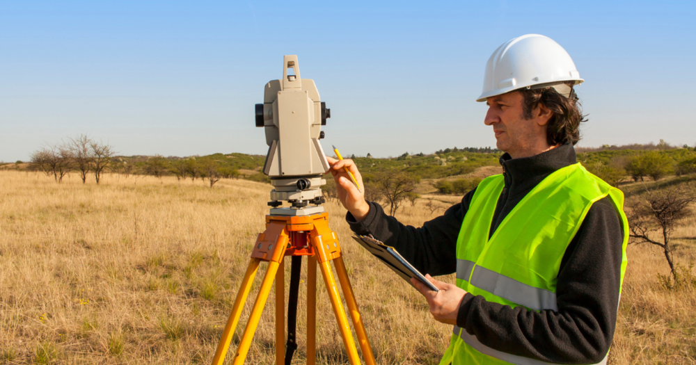 A man in a yellow vest fine tuning a survey instrument on an orange survey tripod.